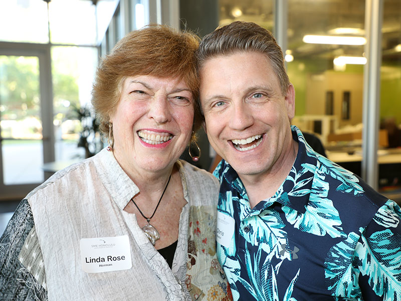 Linda Rose Herrmann smiling with male attendee at SME Honolulu SPOY networking event in Waikiki Honolulu Hawaii, wearing business and aloha attire, engaging with local business leaders indoors