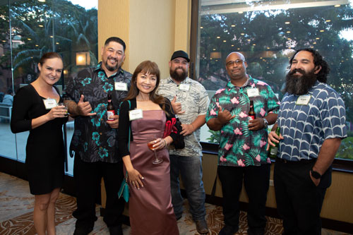Anna Covert and Tharam Singh with professionals holding drinks at SME Honolulu SPOY networking banquet event in Waikiki Honolulu Hawaii, wearing aloha and business attire, making shaka gestures and engaging with local business leaders